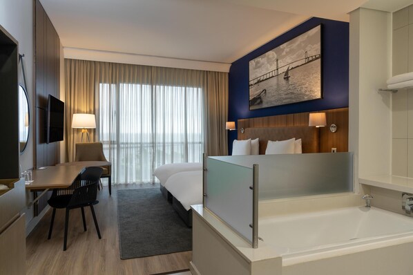 A hotel room with a bed, a desk with a chair, a TV, a painting on the wall, a window with curtains, and a bathtub.