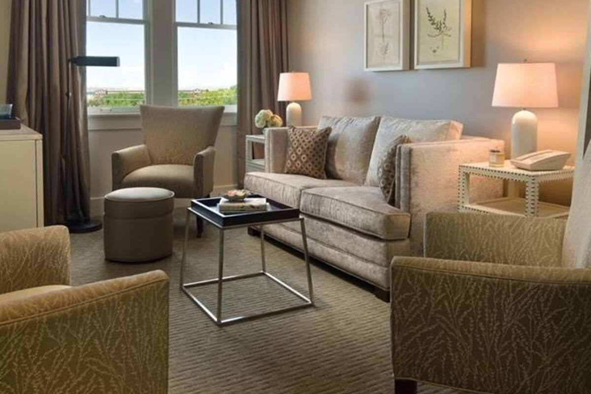 A hotel room with a large window, a sofa, two armchairs, a coffee table, and a side table with lamps.
