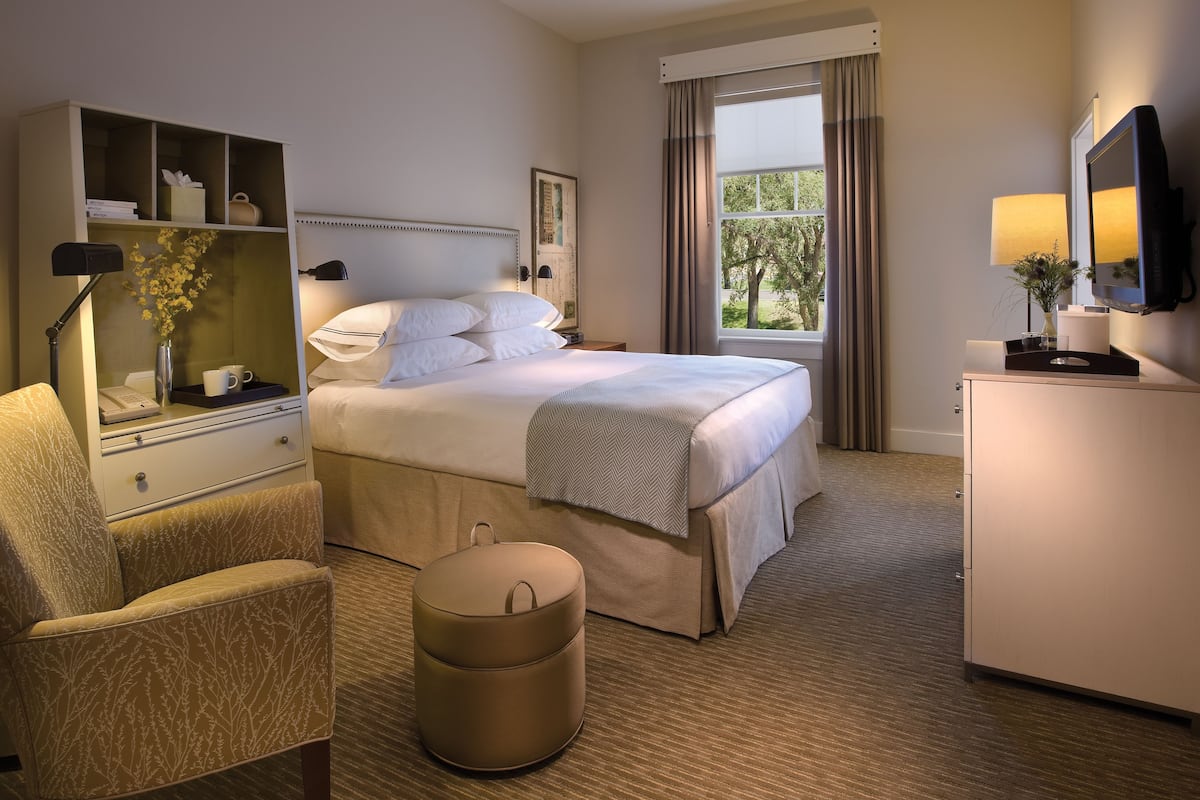 A hotel room with a bed, a chair, a desk with a lamp, a small round ottoman, a dresser, and a television.