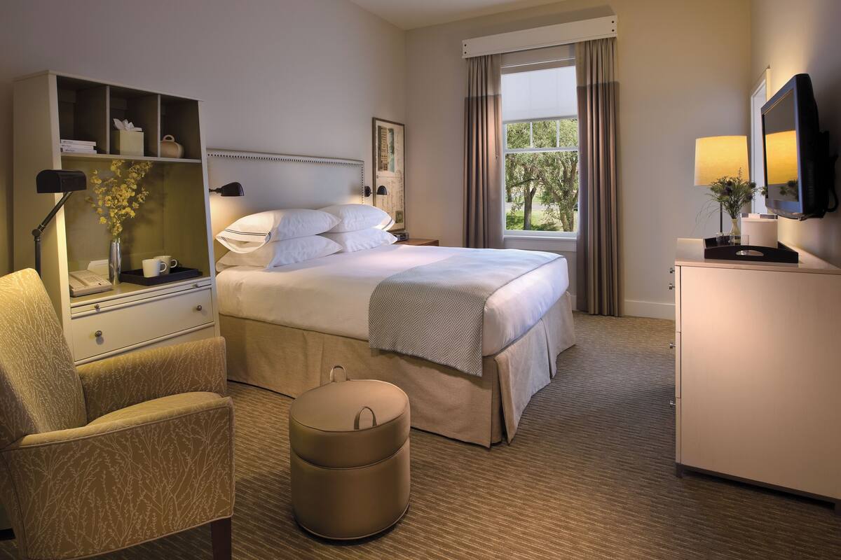 A hotel room with a bed, a chair, a desk with a lamp, a small round ottoman, a dresser, and a television.