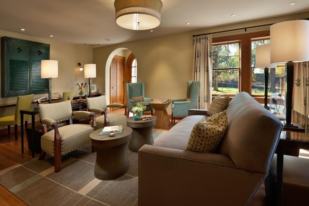 A well-lit living room with a sofa, armchairs, and a coffee table. There are windows with curtains, a fireplace, and a view of the outdoors.