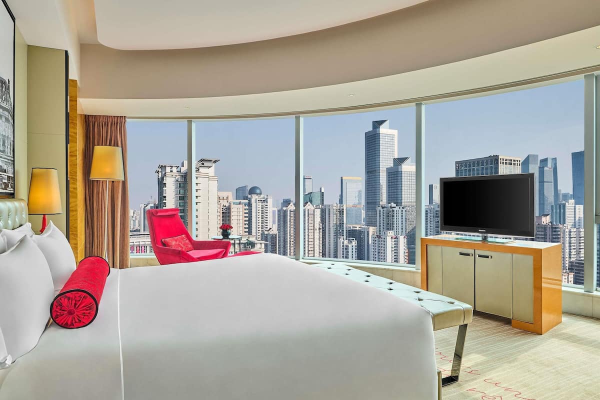 A hotel room with a large bed, a red chair, a TV, and a view of a city skyline through large windows.