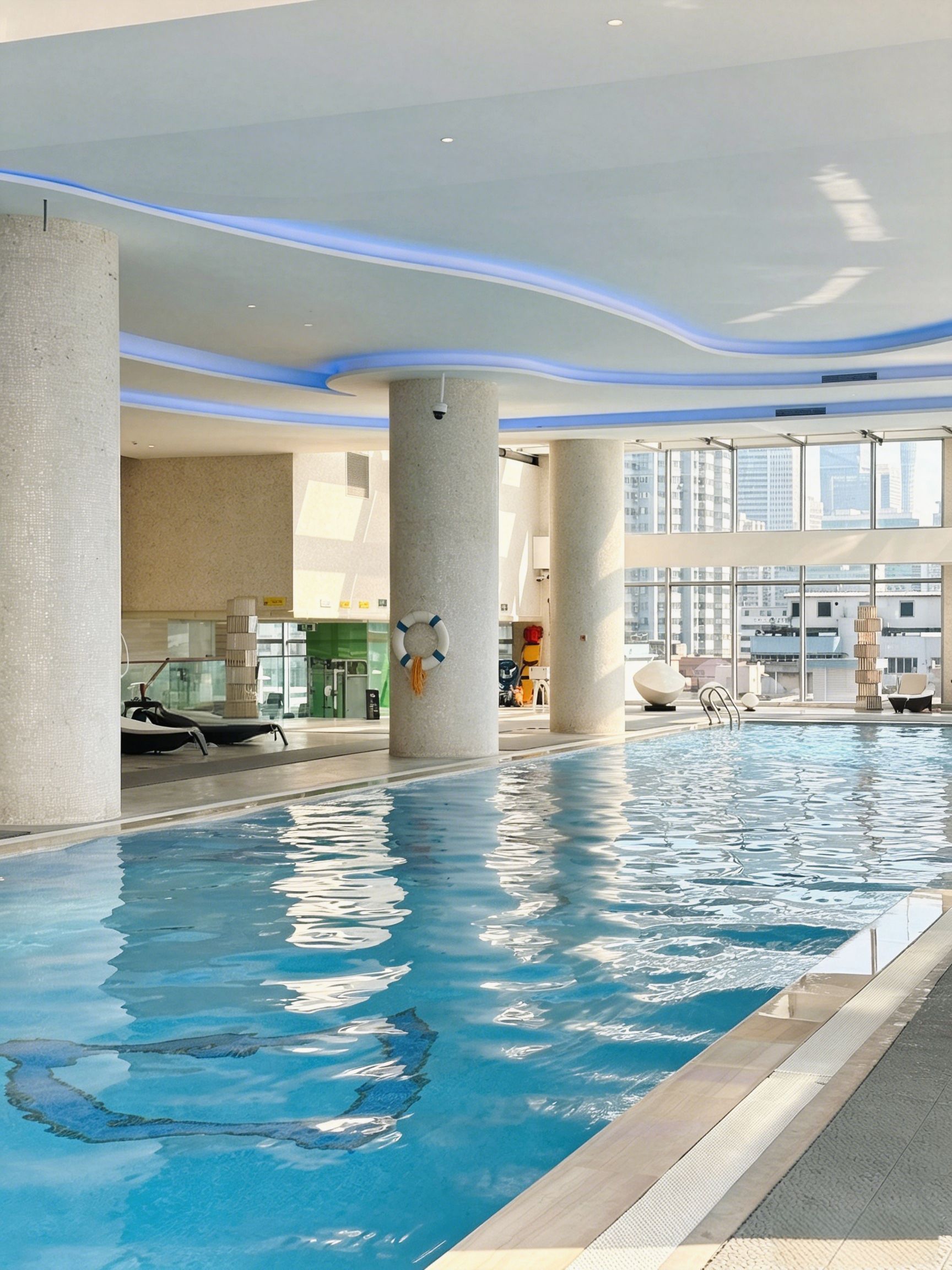 An indoor swimming pool with clear blue water, surrounded by large windows offering views of the cityscape.
