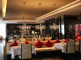 A restaurant with a long dining table, white tablecloths, red cushions, and hanging chandeliers.