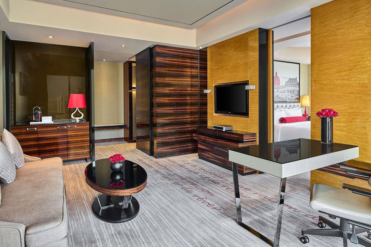 A modern hotel room with a sofa, a coffee table, a TV mounted on a wooden panel wall, and a bed with a cityscape view.
