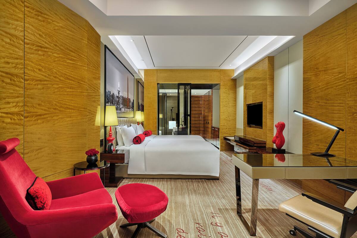 A modern hotel room with a large bed, a desk, a red armchair, and a small round table with a vase of flowers.