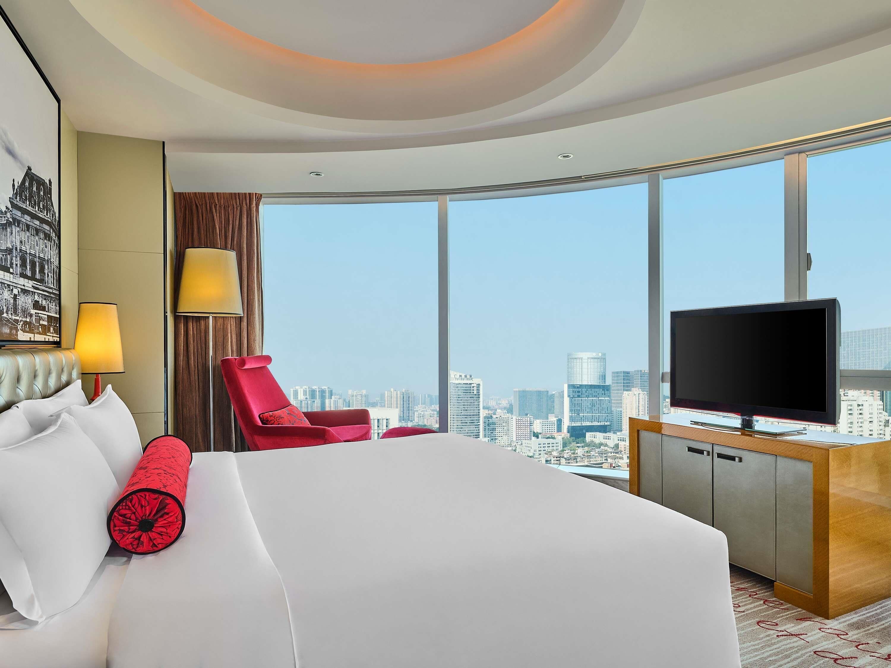 Habitación de hotel con una cama grande, televisión, una silla roja y vistas a la ciudad.