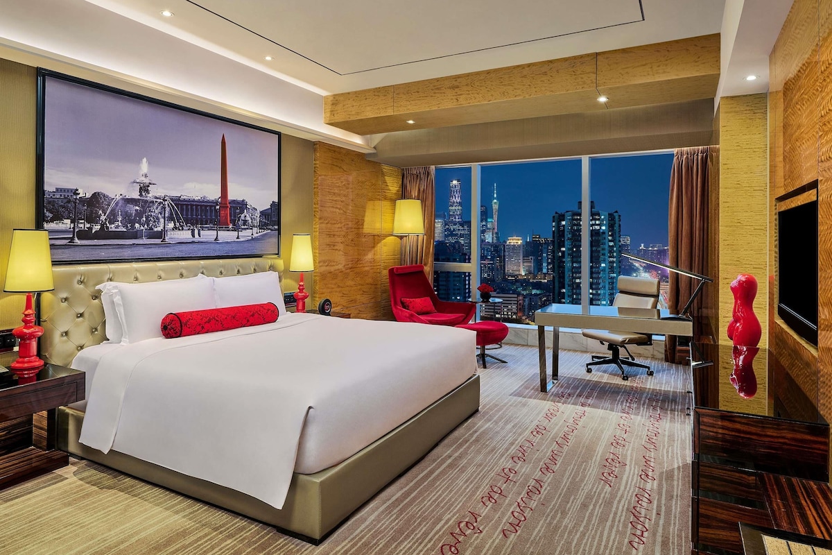 A hotel room with a large bed, a desk, a chair, a TV, and a city view.