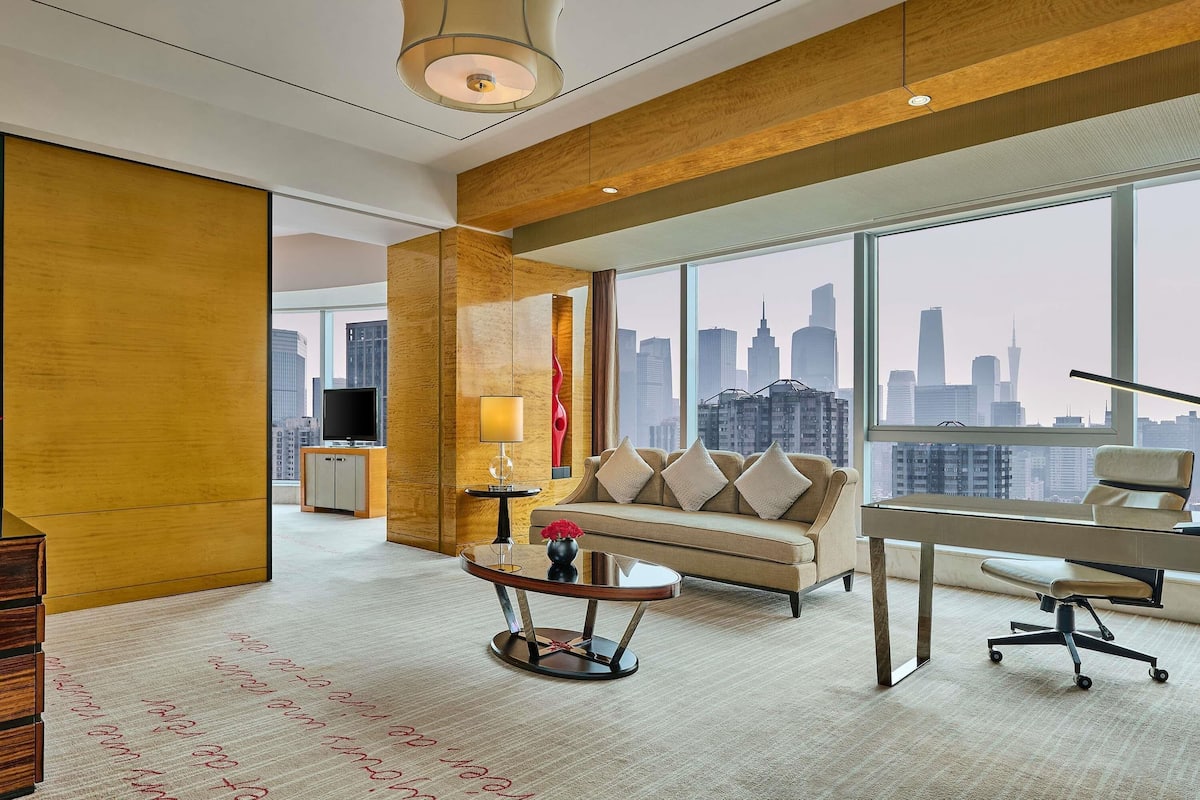A modern hotel room with a city view, a sofa, a table, and a lamp.