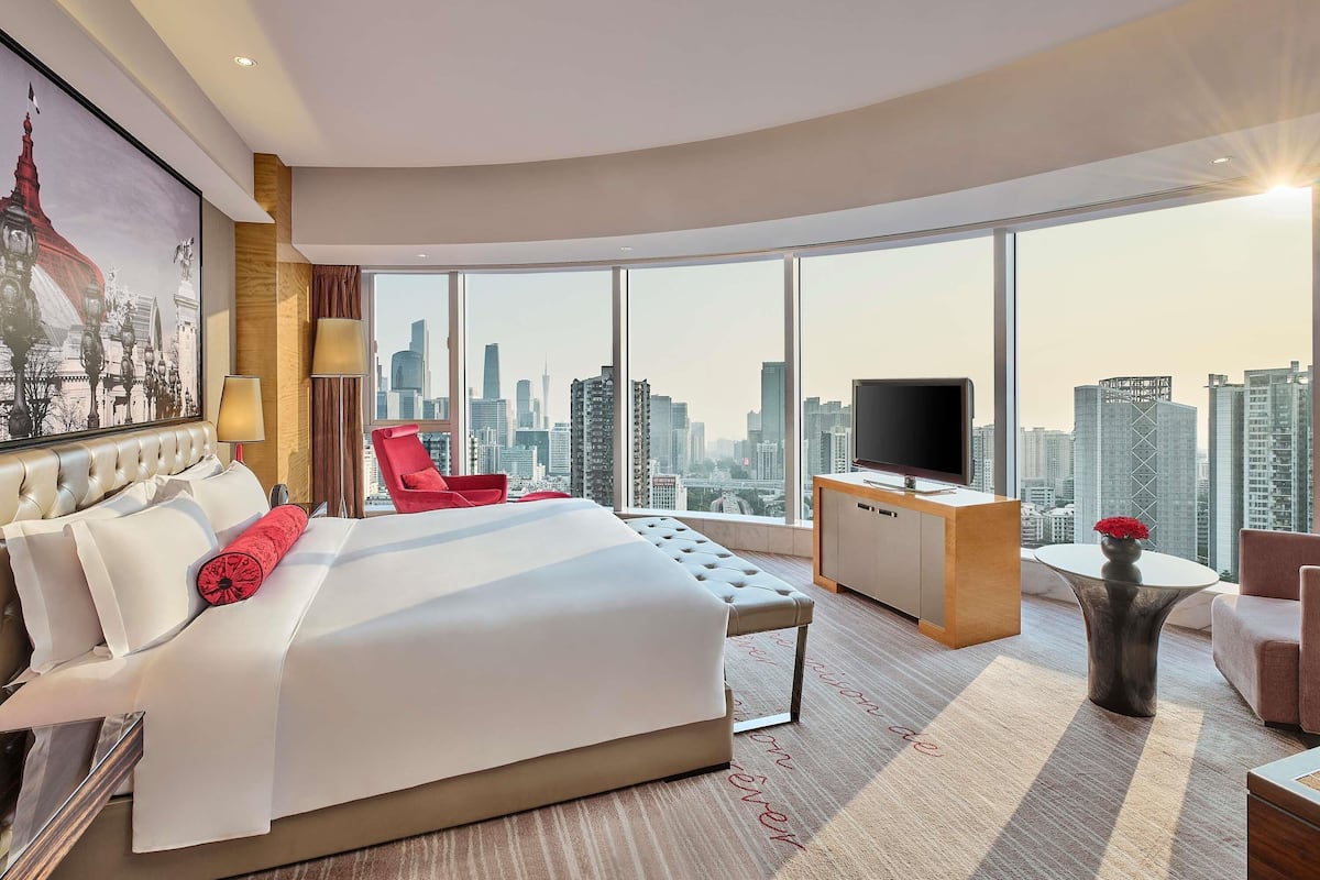A modern hotel room with a large bed, a red chair, a TV, and a city view.