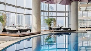 Una piscina interior moderna con paredes de vidrio transparente, sillones de descanso y vistas a la silueta urbana.