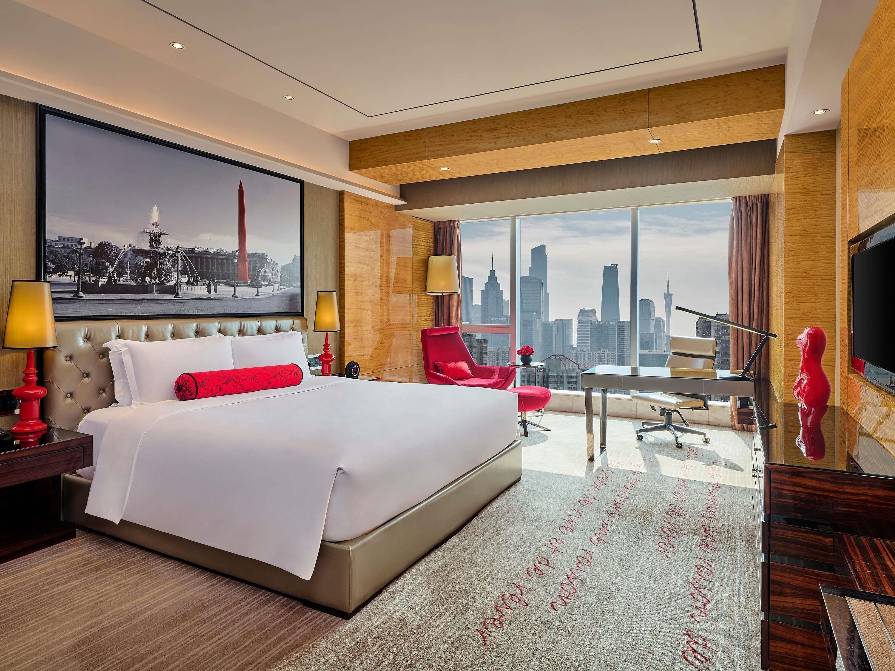 A hotel room with a large bed, a cityscape view, a red chair, and a TV.