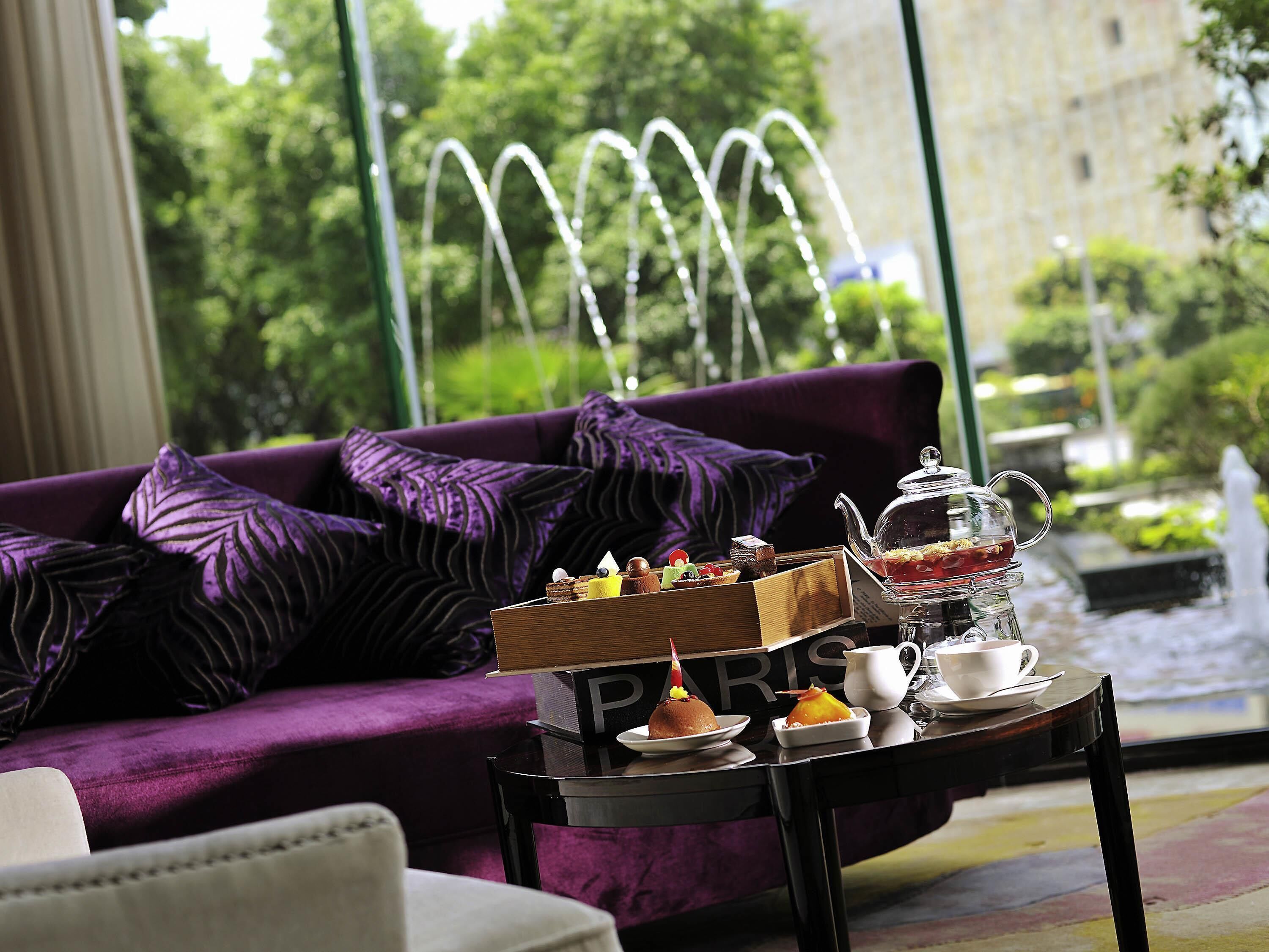 Una habitación con un sofá morado, una mesa con un calentador de agua, una bandeja de postres y vistas a una fuente y vegetación al exterior.