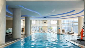 An indoor swimming pool with clear blue water, surrounded by lounge chairs and sunbeds, with large windows offering a view of the cityscape.