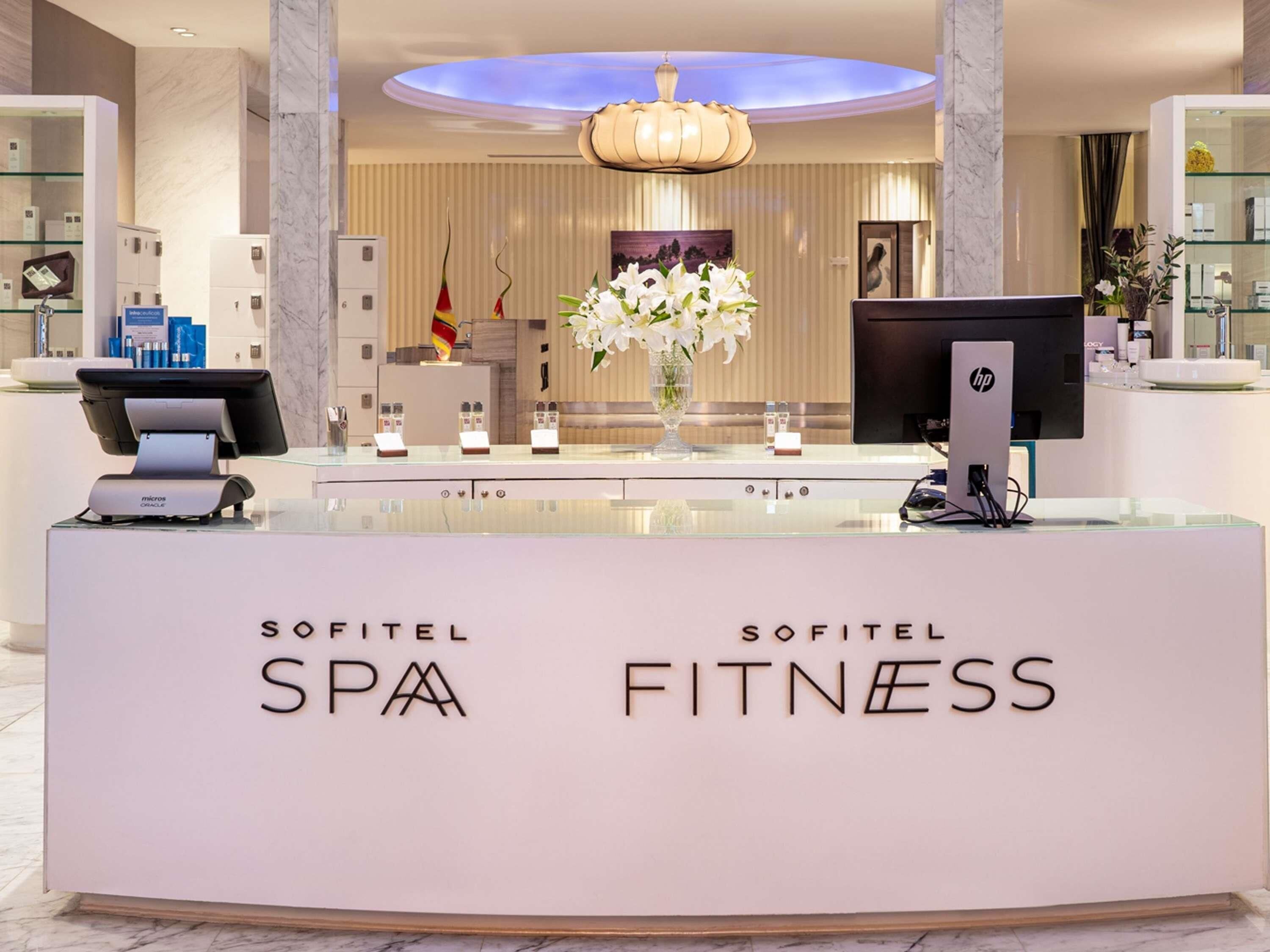 A reception desk with 'Sofitel SPA FITNESS' branding, a modern chandelier, and a vase of white flowers.