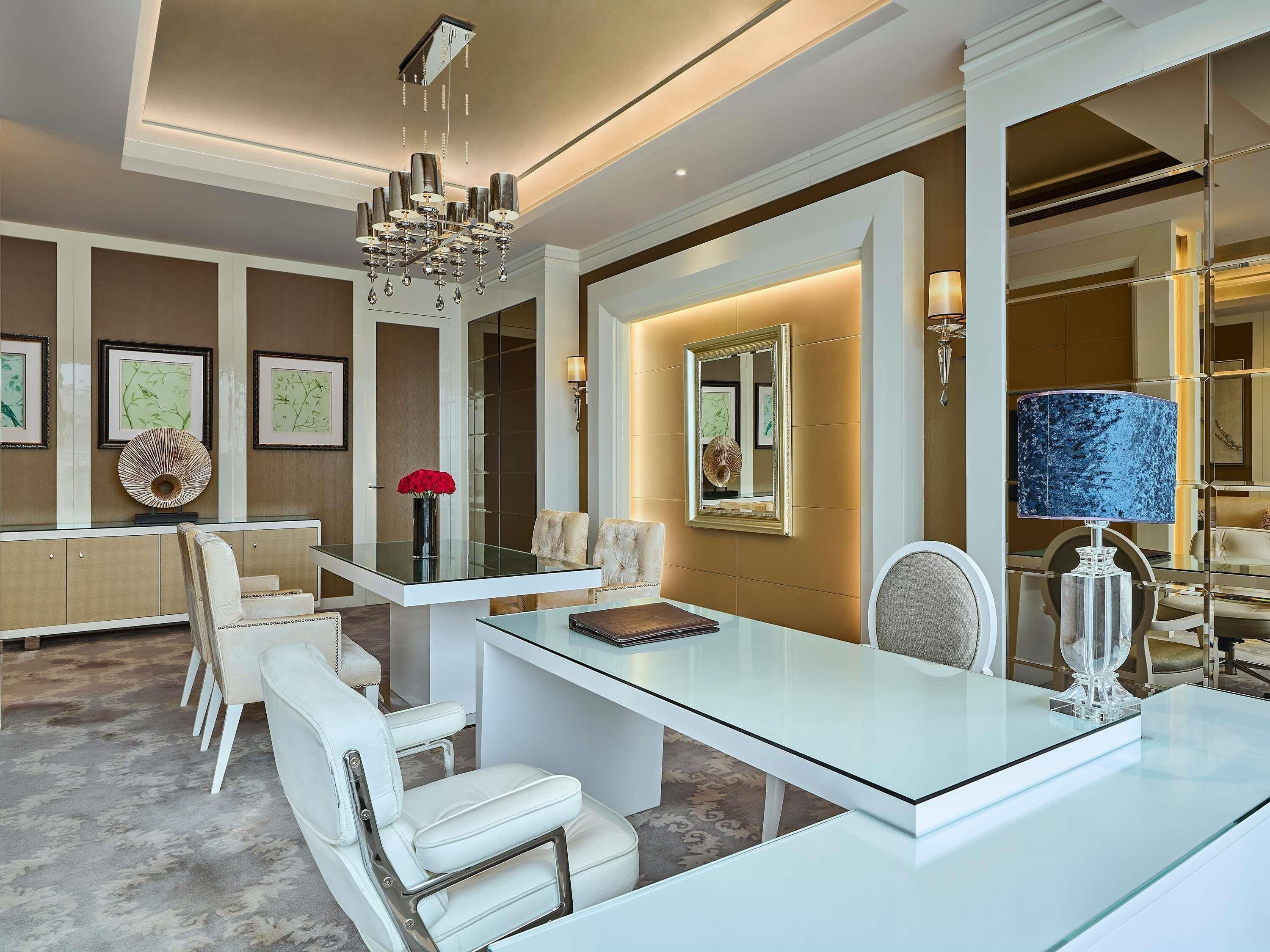 A modern interior with a glass coffee table, white leather chairs, a chandelier, framed artwork, and a mirror.