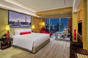 Camera d'albergo con un letto grande, una scrivania con lampada, una sedia, una televisione e vista sullo skyline della città.