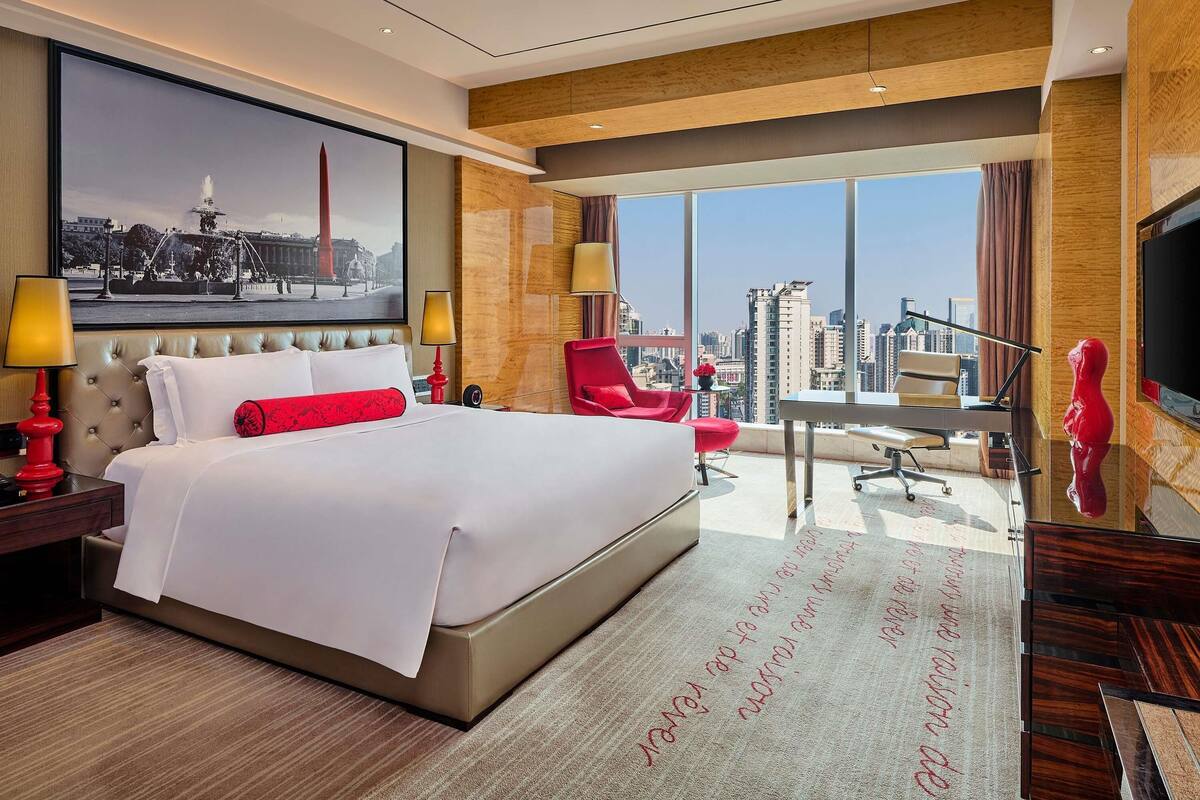A hotel room with a large bed, a desk, a red chair, a TV, and a city view.