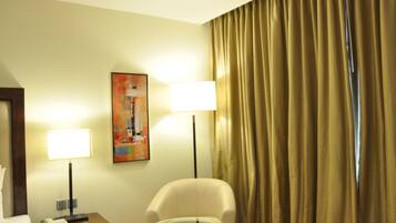 A hotel room with a bed, a nightstand with a lamp, a glass table, a chair, and a painting on the wall.