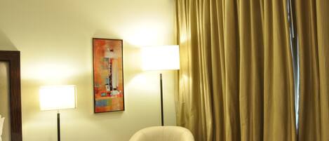 Una habitación de hotel con una cama, una mesita de noche con lámpara, una mesa de vidrio, una silla y un cuadro en la pared.
