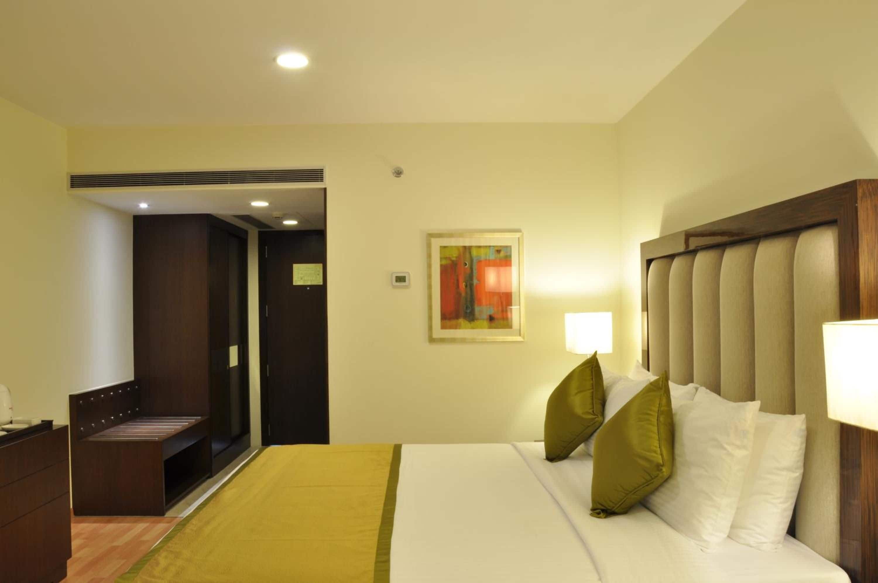 Habitación de hotel con cama, mesita de noche, escritorio, silla, puerta y lámpara fijada a la pared.