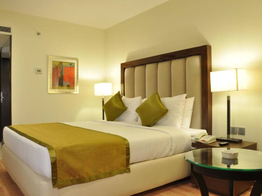 Habitación de hotel con cama, dos lámparas de noche, una mesita de vidrio y un cuadro en la pared.