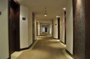 A hotel hallway with carpeted floors, doors on both sides, and a number sign on the wall. - Adarsh Hamilton (Bengaluru)