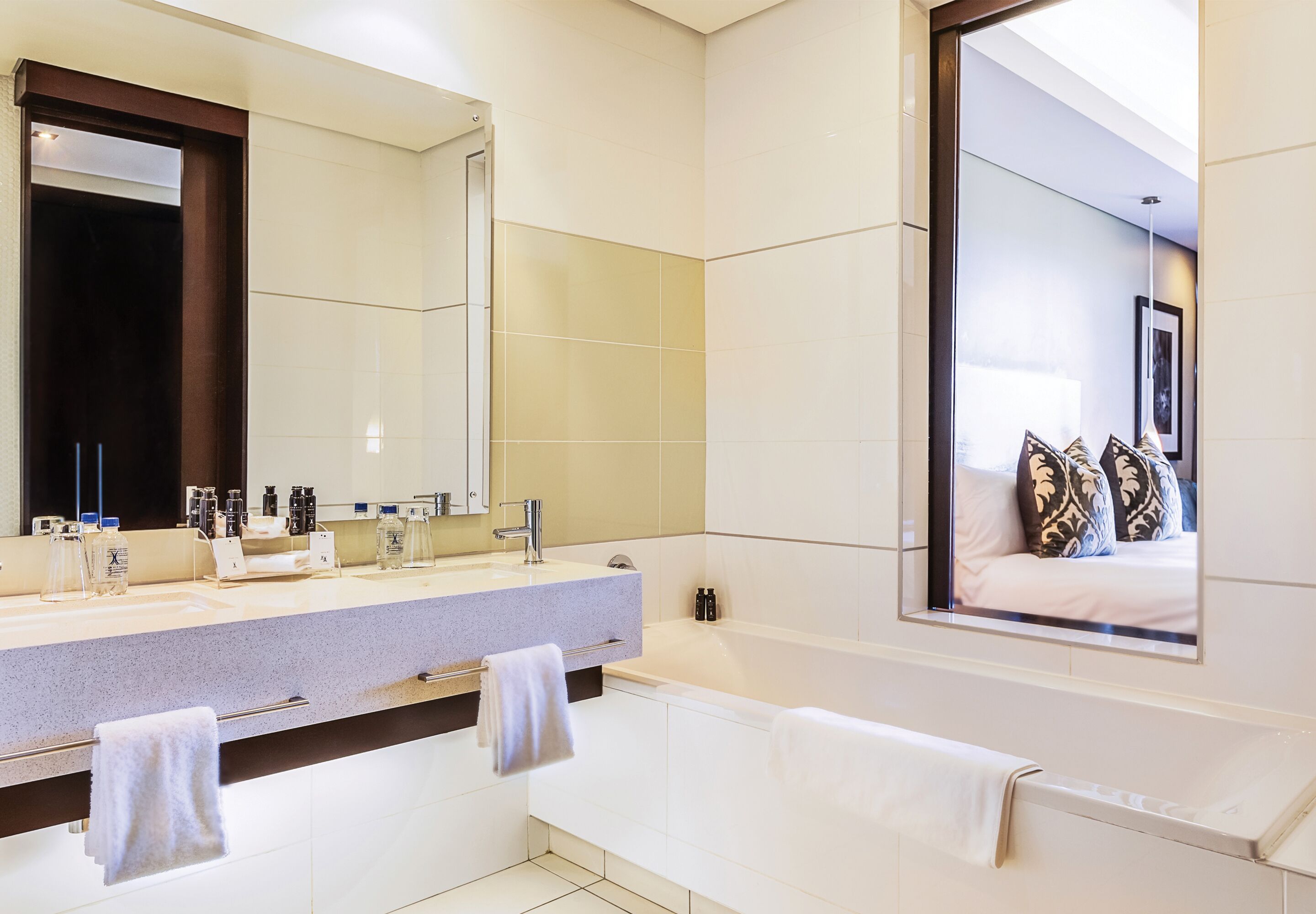 A modern bathroom with a large mirror, two sinks, a bathtub, and a window with a view of a bedroom.