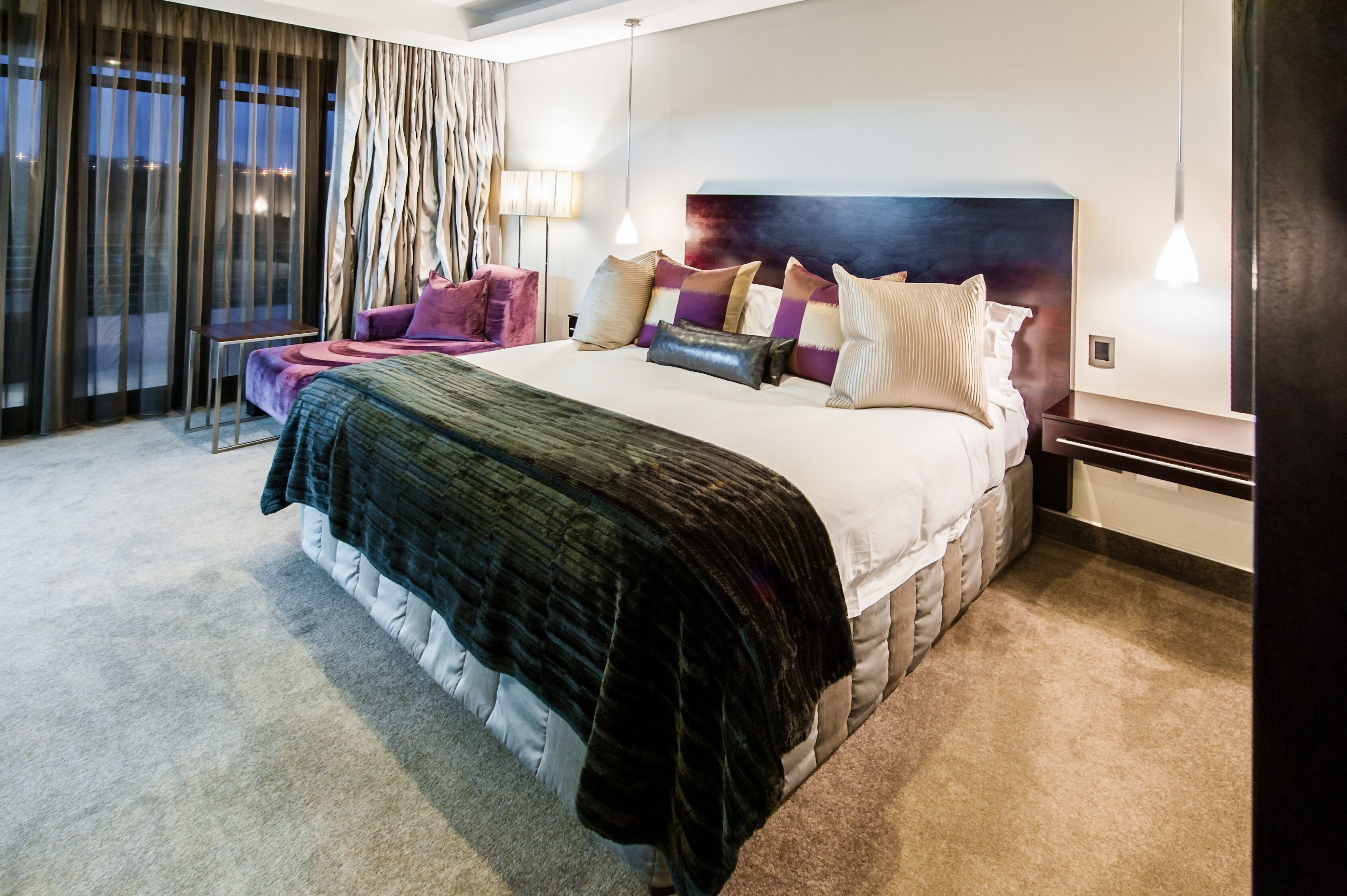 A modern hotel room with a large bed, a side table, a chair, and a view of the city at dusk.