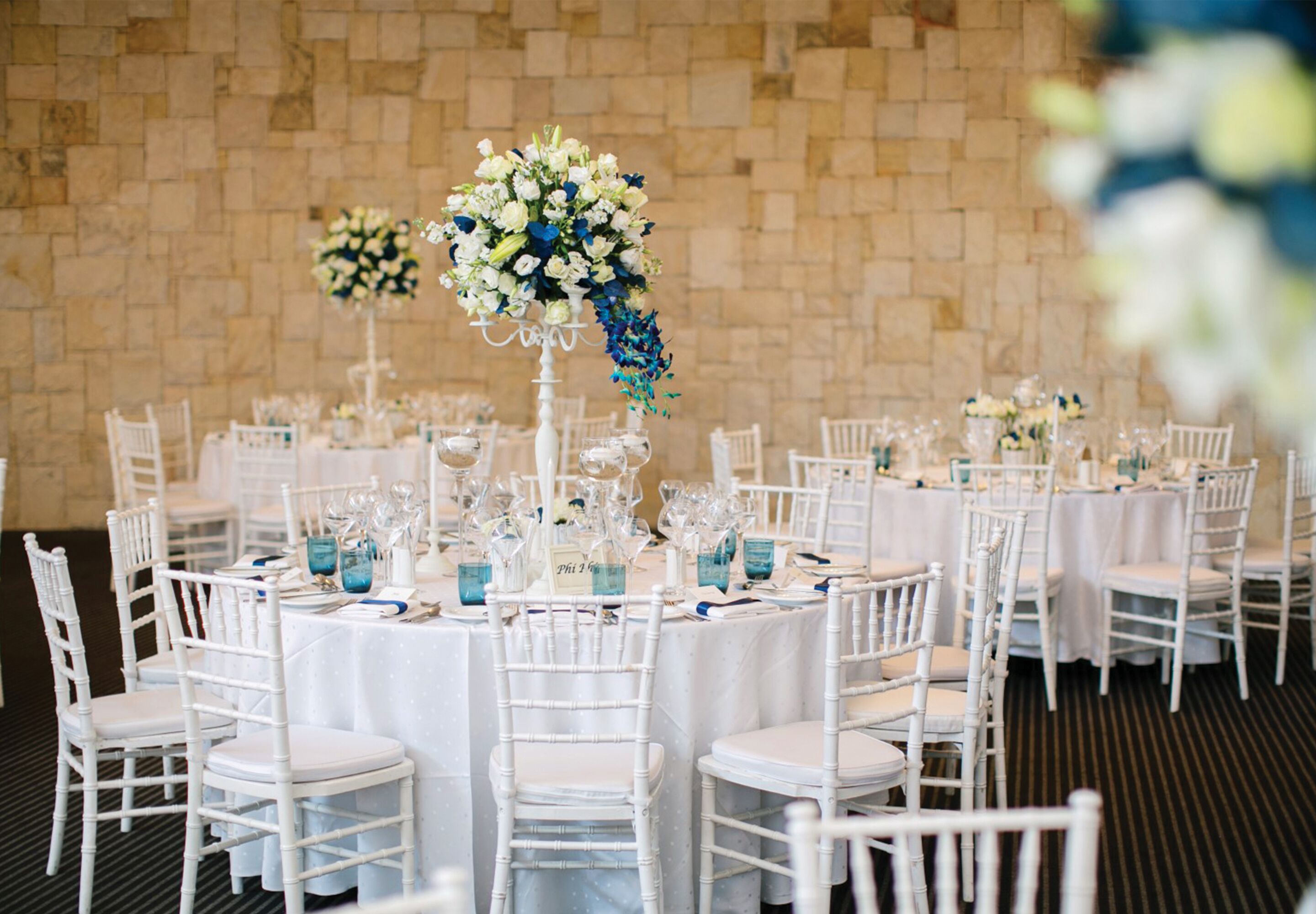 Un salón de banquetes con mesas redondas dispuestas para un evento formal, sillas blancas y centros de mesa florales.