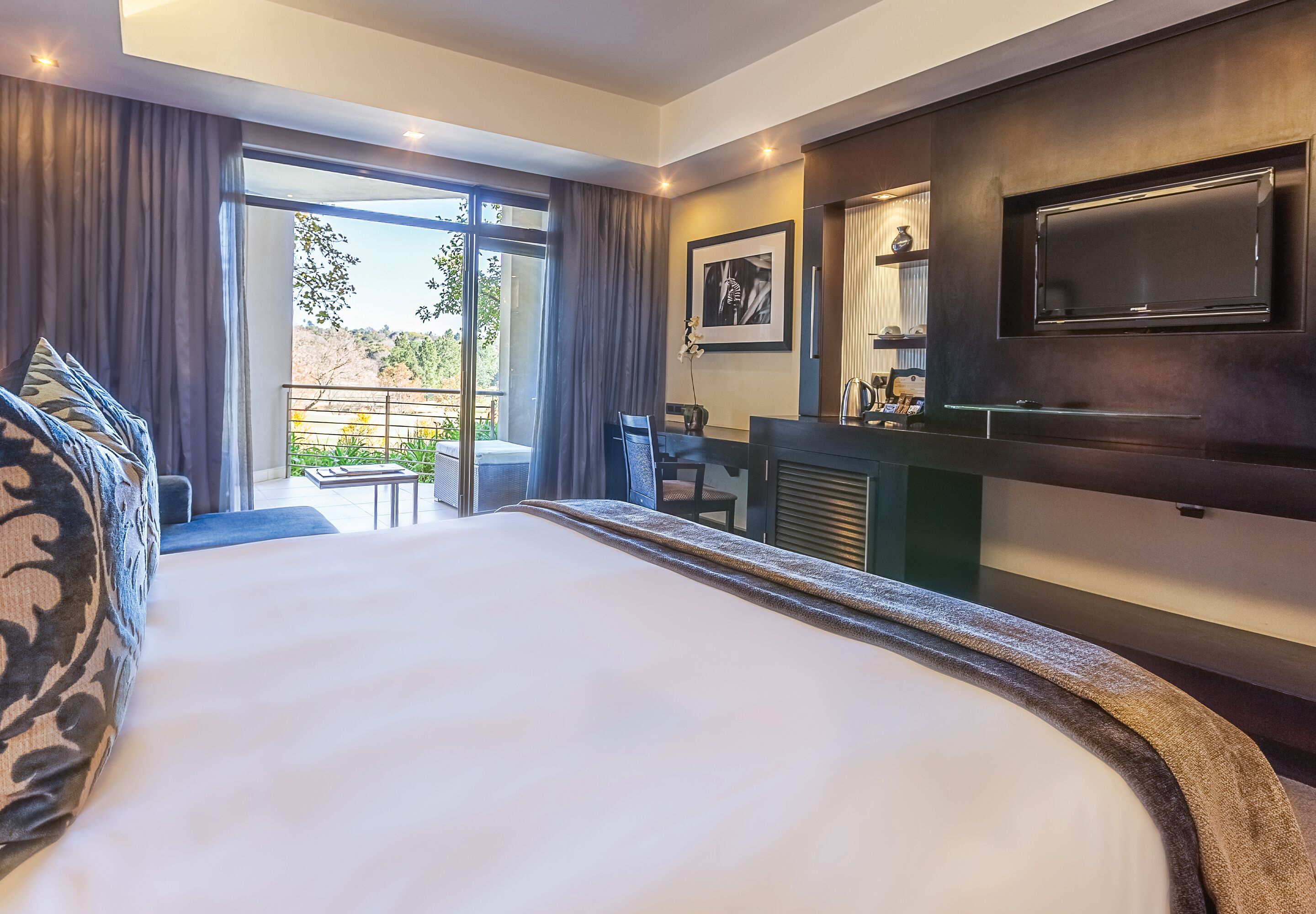 A hotel room with a large bed, a TV mounted on the wall, a desk with a chair, and a view of the outdoors through a sliding door.