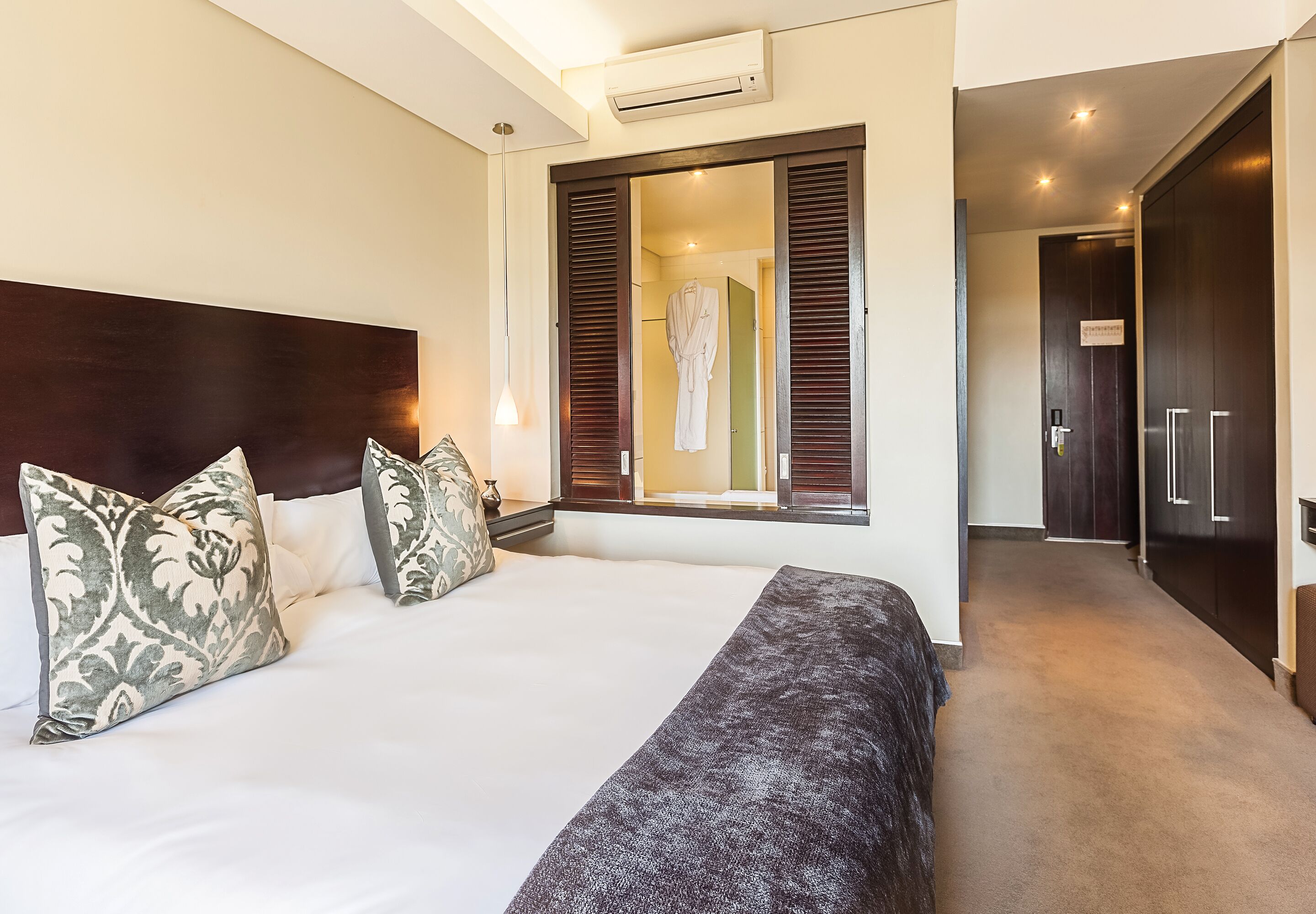 A hotel room with a large bed, a window with wooden shutters, a robe hanging on the wall, and a dark wooden wardrobe.