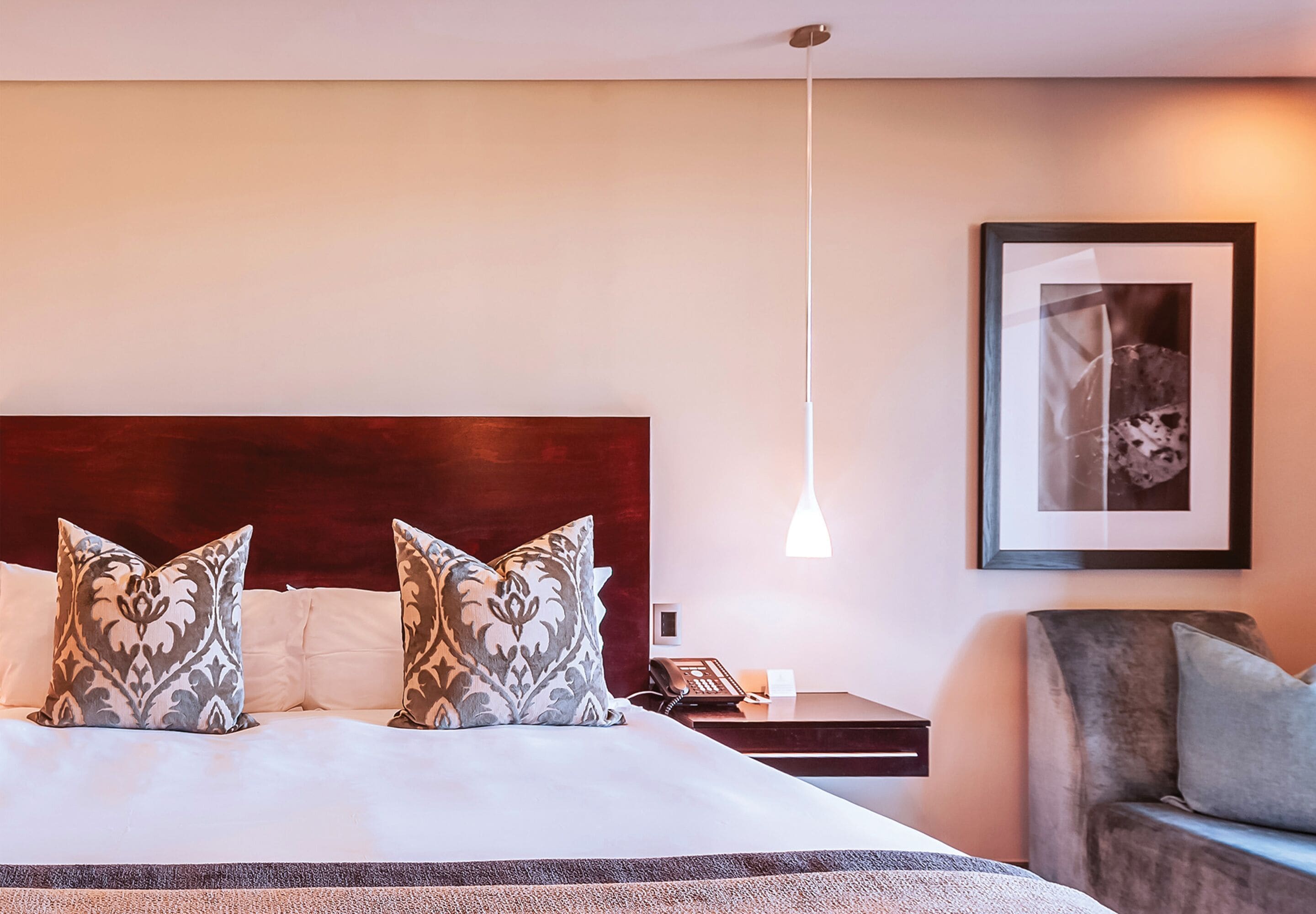 A hotel room with a large bed, two patterned pillows, a bedside table with a phone, a framed picture on the wall, and a hanging light fixture.