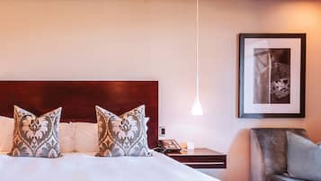 Una habitación de hotel con una cama grande, dos almohadas estampadas, una mesita de noche con un teléfono, un cuadro enmarcado en la pared y una lámpara colgante.