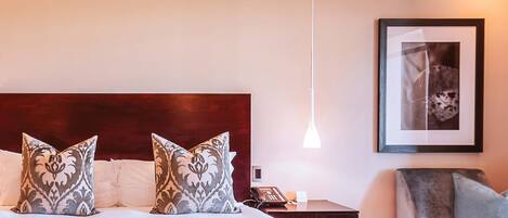 A hotel room with a large bed, two patterned pillows, a bedside table with a phone, a framed picture on the wall, and a hanging light fixture.