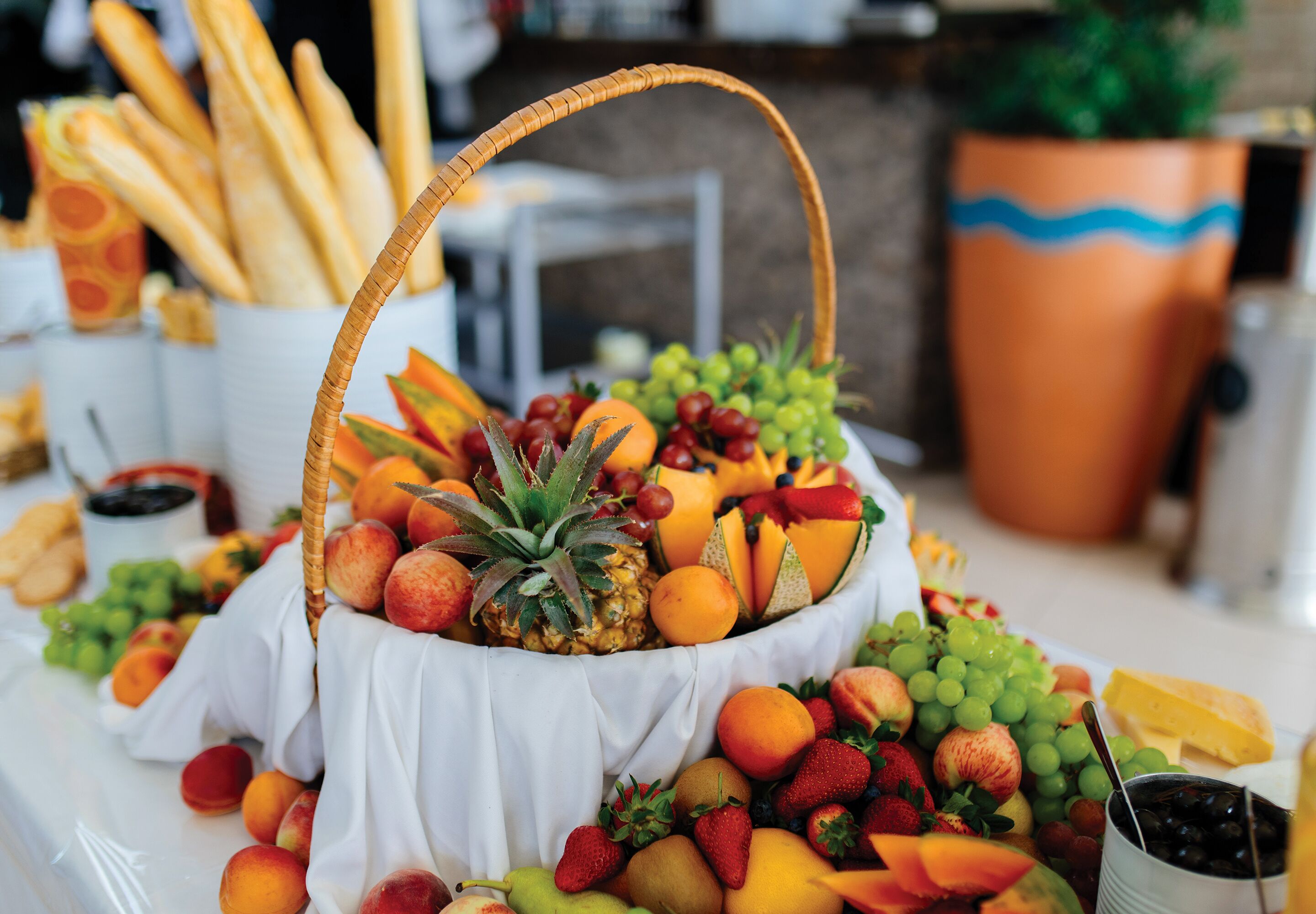 A fruit and cheese platter with a wicker basket, surrounded by baguettes and a potted plant.