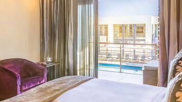 Habitación de hotel con una cama, un sillón púrpura, una mesita con una taza y vistas a una piscina y un edificio.