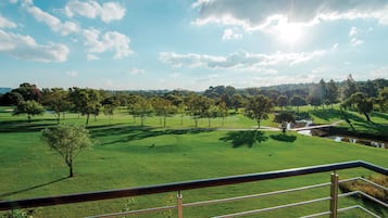 Vista de um campo de golfe, com céu claro e um corpo de água ao longe.
