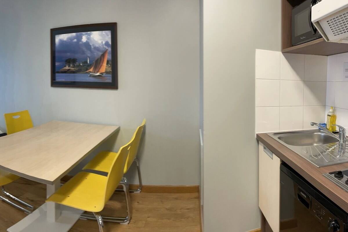 Une chambre d’hôtel compacte comprenant un lit, un petit bureau, une table à manger avec des chaises jaunes, un coin cuisine et une salle de bain avec un miroir et un lavabo.