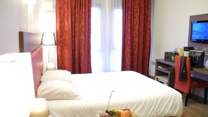 A hotel room with a bed, a television, a desk with a lamp, a basket of fruit, and red curtains. - Villa Val Sénart (Quincy-sous-Senart)