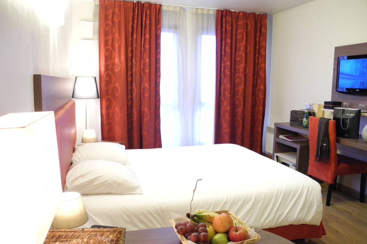 Une chambre d’hôtel avec un lit, une télévision, un bureau avec une lampe, un panier de fruits et des rideaux rouges.