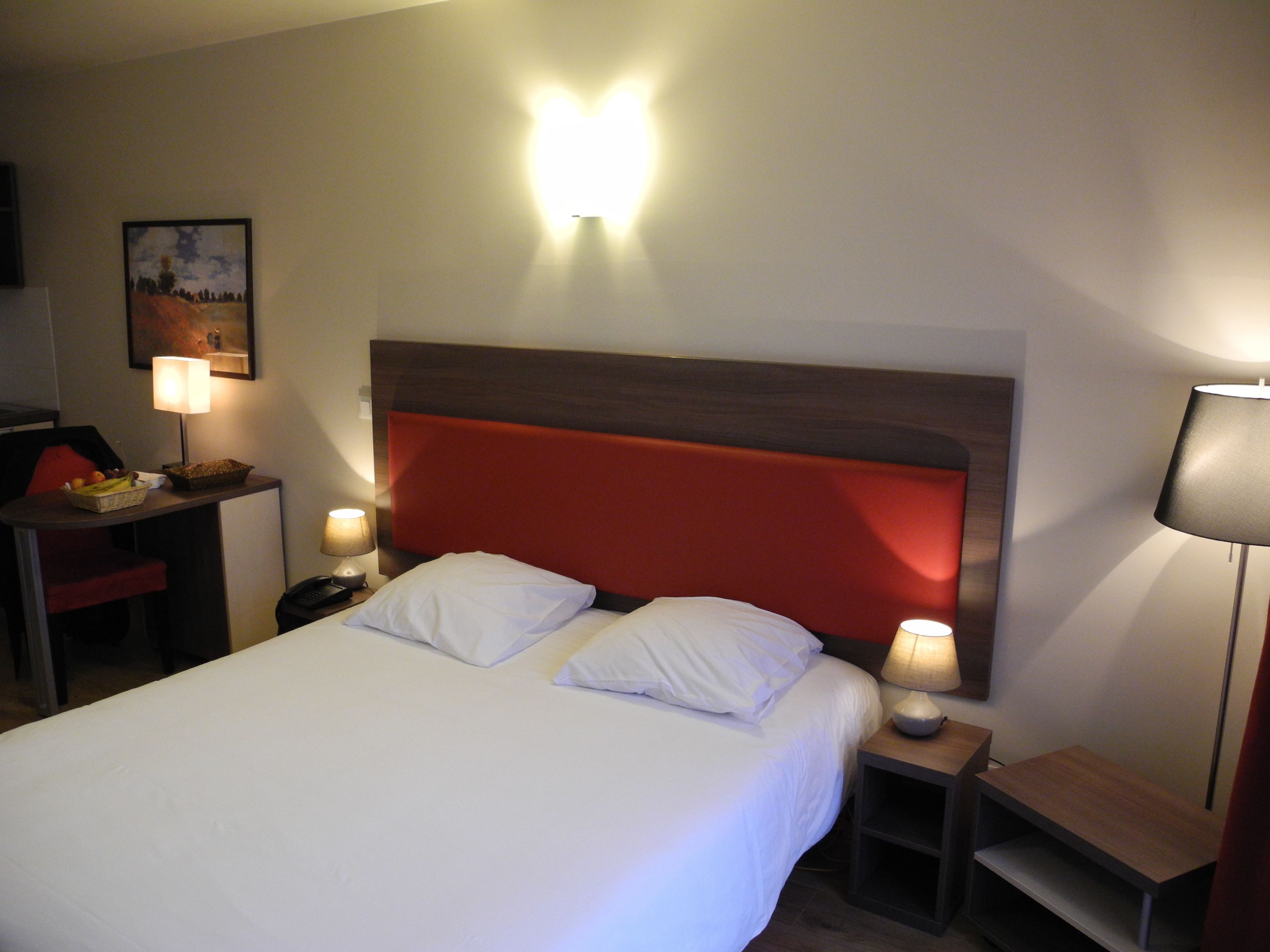 Une chambre d’hôtel comprenant un lit, des lampes de chevet, une table de chevet, une chaise, une petite table avec un panier et une lampe fixée au mur.