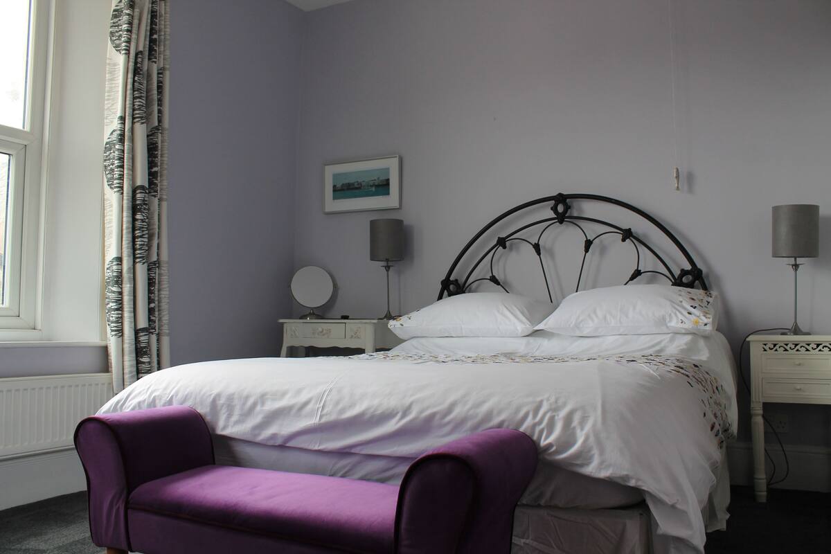 A bedroom with a bed, a nightstand, a lamp, a chair, and a window with curtains.