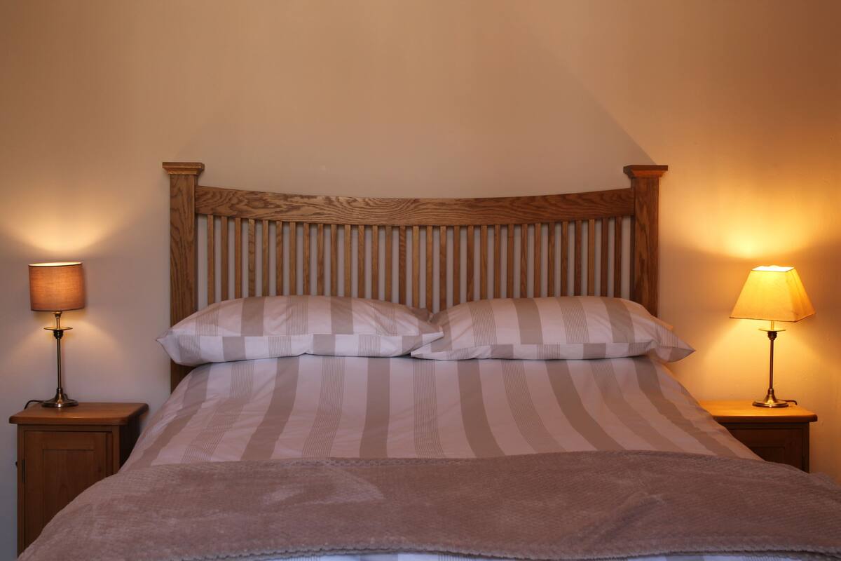 A neatly made bed with striped bedding and two pillows, flanked by two bedside tables with lamps, set against a plain wall.