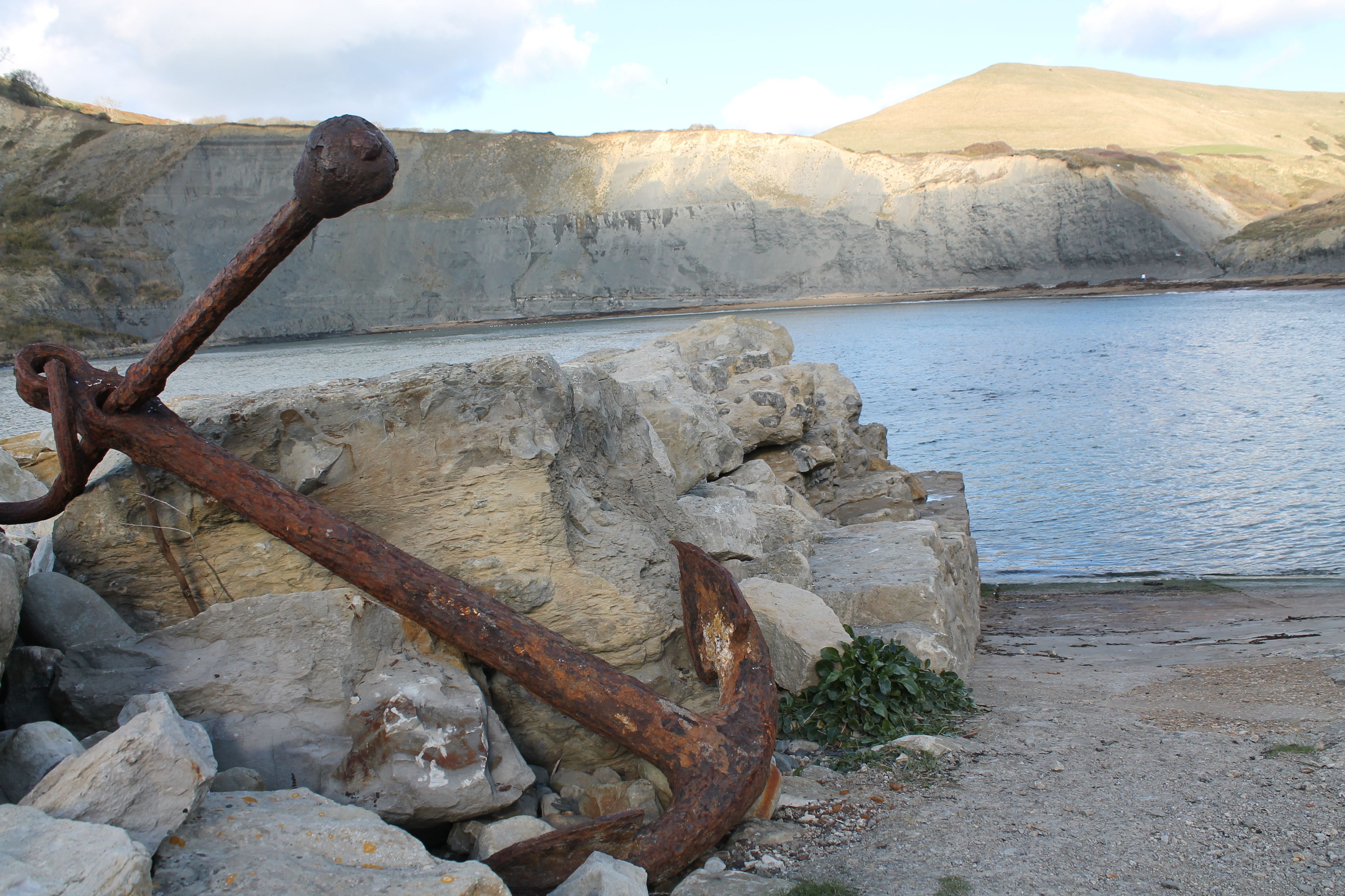 A rusty anchor on rocks by a calm body of water with cliffs in the background.