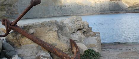 A rusty anchor on rocks by a calm body of water with cliffs in the background.