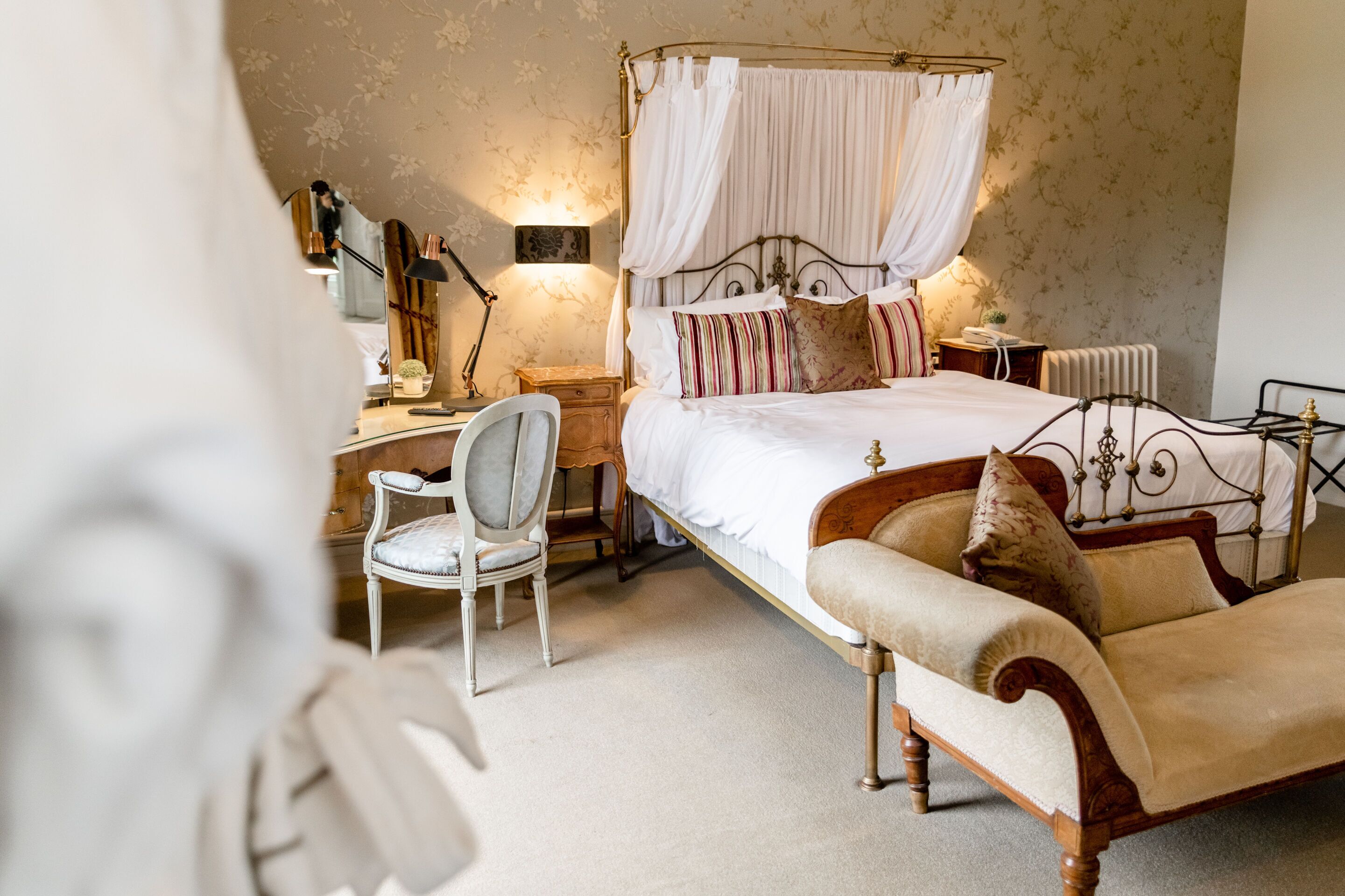 A bedroom with a four-poster bed, a bedside table with a lamp, a chair, and a radiator.