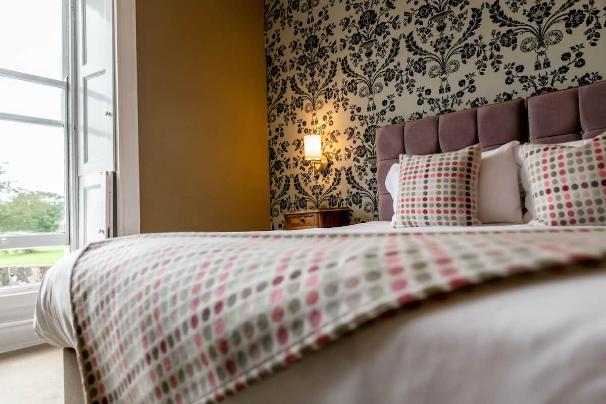 A bedroom with a bed, a nightstand, a wall-mounted lamp, and a window.