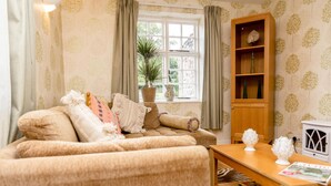 A cozy living room with a sofa, a wooden side table, a bookshelf, and a window with curtains. - Sweeney Hall Hotel (Oswestry)
