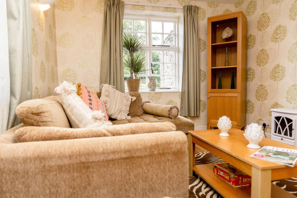 A cozy living room with a sofa, a wooden side table, a bookshelf, and a window with curtains.